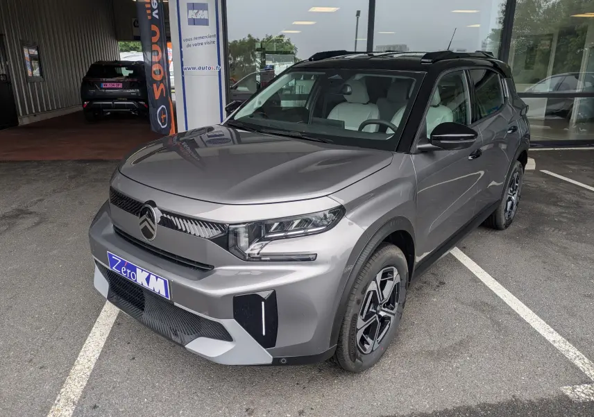 Vue 3/4 avant droite d'une Citroën C3 Aircross gris Mercuy avec toit noir, garée devant un bâtiment vitré.