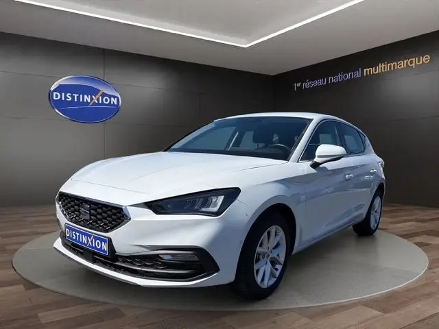 Vue 3/4 avant d'une SEAT Leon blanche 2025, avec calandre noire et phares LED, sur podium intérieur showroom.
