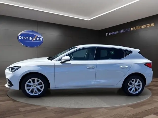 Vue de profil côté gauche d'une SEAT Leon blanche 2025 dans un showroom avec éclairage moderne et sol en bois.