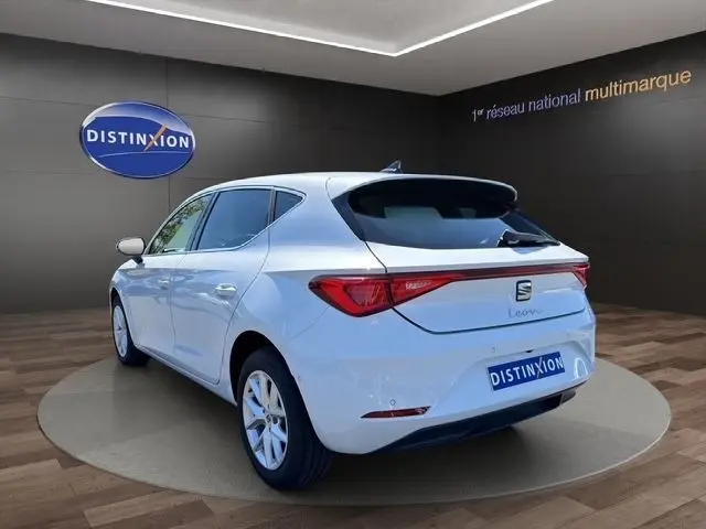 Vue 3/4 arrière gauche d'une SEAT Leon blanche 2025 dans un showroom avec logo Distinxion en arrière-plan.