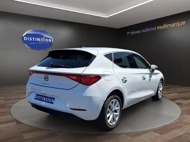Vue 3/4 arrière droite d'une SEAT Leon blanche 2025 dans un showroom avec logo Distinxion en arrière-plan.