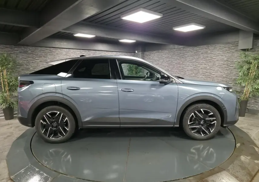 Profil droit d’un Peugeot 3008 hybride 2025 bleu métallisé avec jantes alliage noires et toit noir dans un showroom.