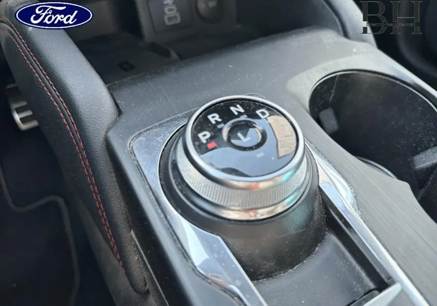 Gros plan sur la molette de sélection de vitesses et l'accoudoir central en cuir noir avec surpiqûres rouges dans un Ford Kuga 2024.