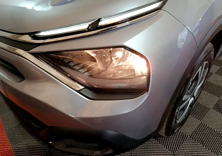 Gros plan sur le phare avant droit allumé d'une Citroën C4 gris foncé, avec partie de la roue visible.