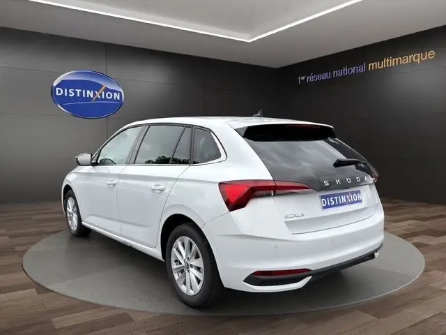 Vue 3/4 arrière droite d'une Skoda Scala blanche 2025 avec feux arrière LED et logo distinctif sur fond showroom.