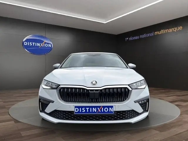 Vue frontale d'une Skoda Scala blanche 2025 avec calandre noire et logo au centre, sur fond intérieur showroom.