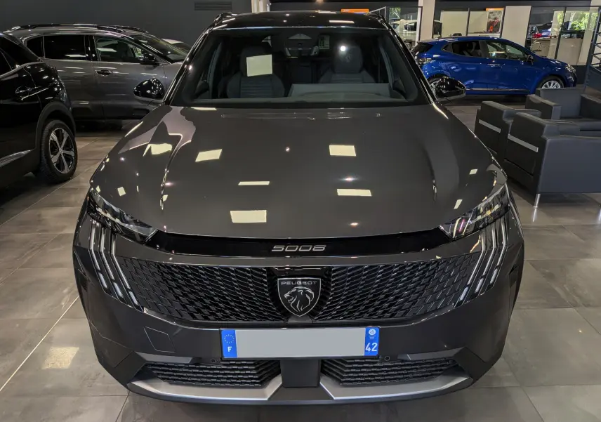 Vue avant d'un Peugeot 5008 Hybrid gris titane avec toit noir, calandre noire et logo lion central visible en showroom.