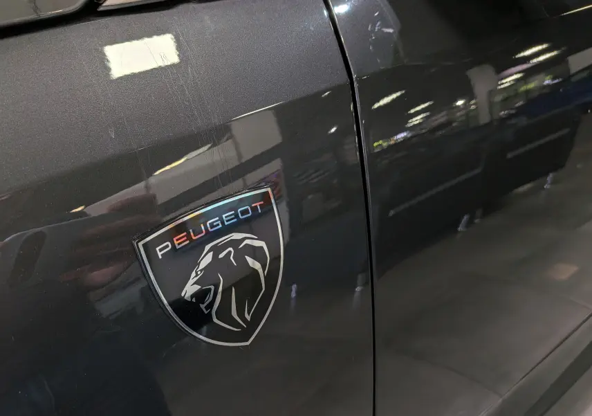 Gros plan sur l’écusson Peugeot noir et gris sur la porte gris titane d’un 5008 hybride 2025.