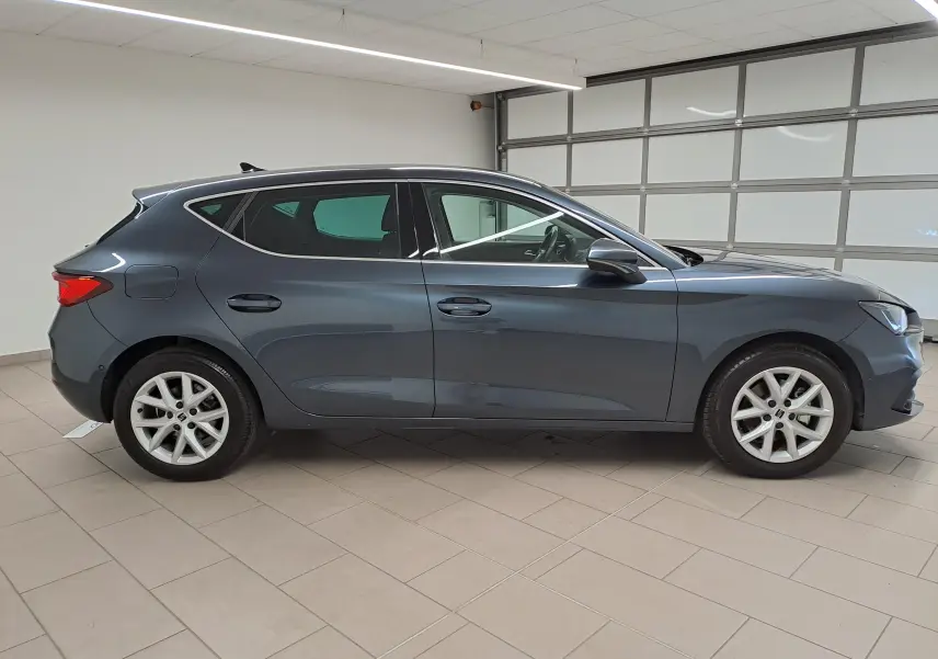 Vue de profil côté gauche d'une SEAT LEON 1.5 eTSI Hybrid gris magnétique avec vitres arrière surteintées en intérieur showroom.