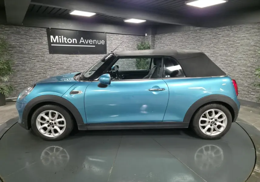 Vue de profil côté gauche d'une MINI Cabriolet bleu avec capote noire et jantes argentées en intérieur showroom.