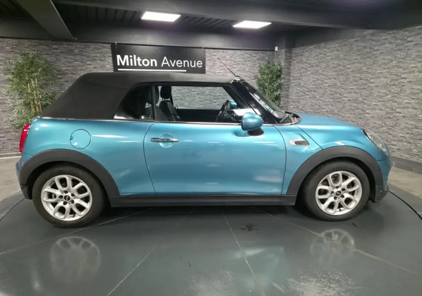 Vue de profil côté gauche d'une MINI Cabriolet bleu avec capote noire et jantes argentées en intérieur showroom.