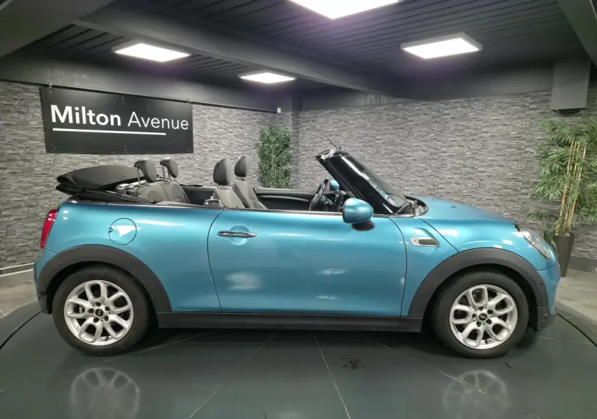 Vue latérale droite d'une MINI Cabriolet bleu avec capote noire repliée et jantes argentées en intérieur showroom.