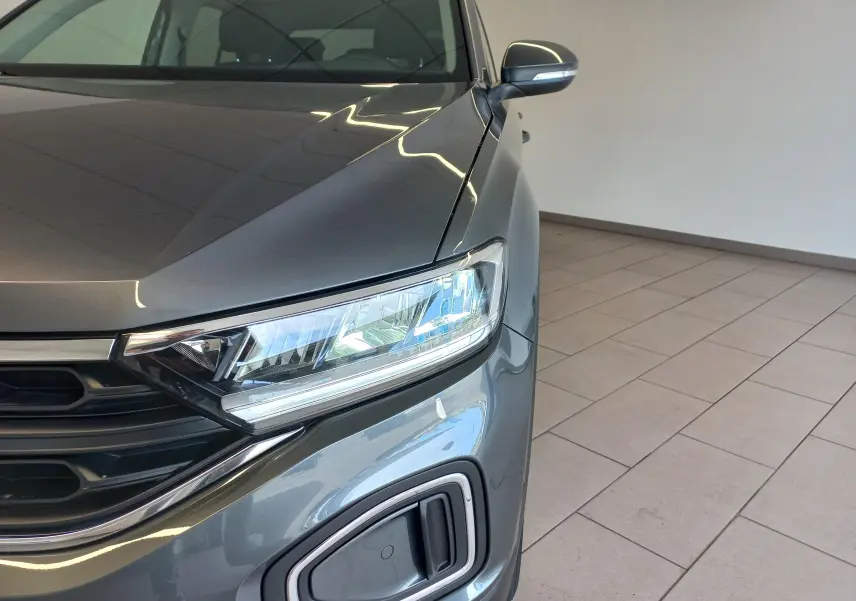 Gros plan sur l'avant droit du Volkswagen T-Roc 2025 gris Indium, mettant en valeur le phare et la calandre moderne.