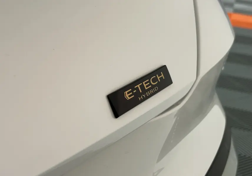 Gros plan sur l'emblème E-TECH Hybrid noir et or sur la carrosserie blanc nacré arrière droite d'une Renault Symbioz.