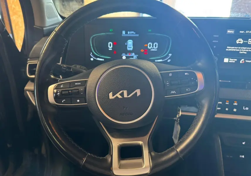 Vue rapprochée du volant noir du Kia Sportage 2022 avec commandes intégrées et tableau de bord numérique éclairé.