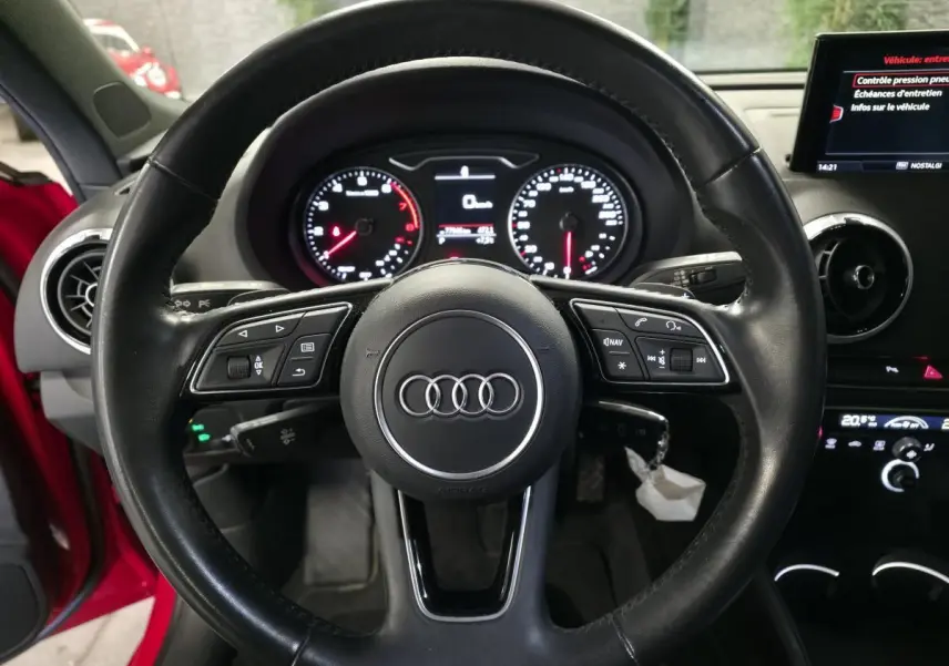Vue intérieure centrée sur le volant noir multifonction de l'Audi A3 Sportback rouge avec tableau de bord et écran central visibles.