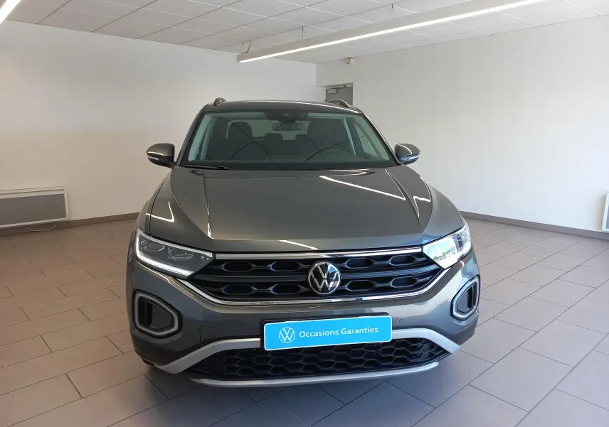 Vue avant d'un Volkswagen T-Roc 2025 gris Indium avec calandre noire et phares LED dans un showroom.