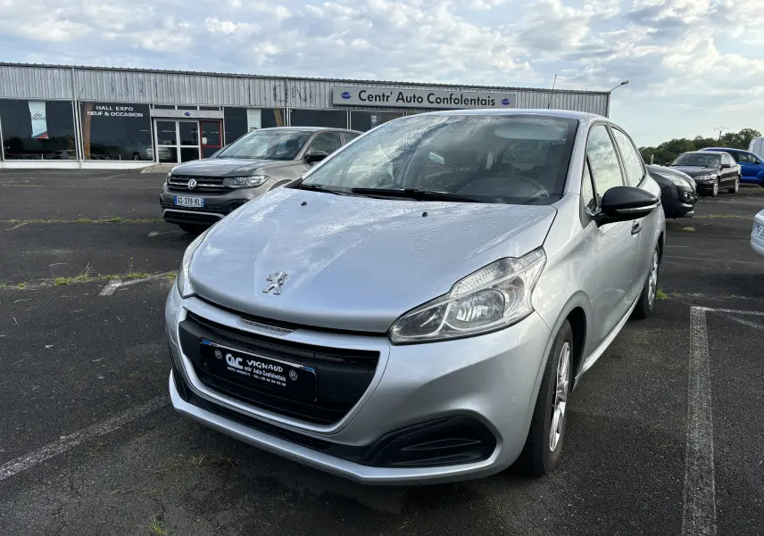 Peugeot 208 gris argent vue 3/4 avant droit, garée sur un parking devant un bâtiment commercial.