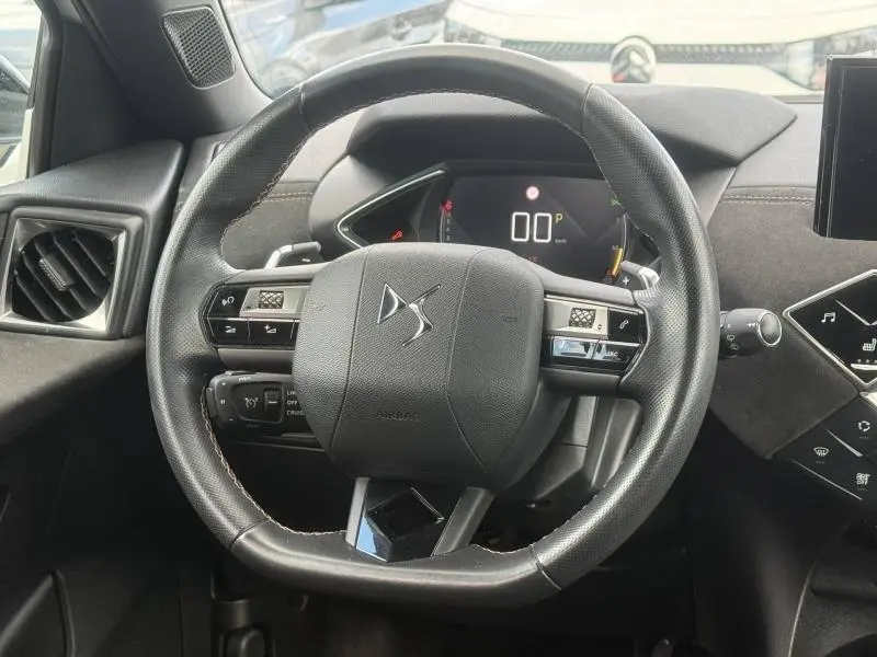 Vue rapprochée du volant cuir noir du DS3 Crossback avec commandes intégrées et tableau de bord digital en arrière-plan.