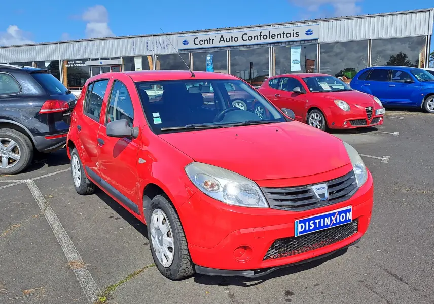 Vue 3/4 avant d'une Dacia Sandero rouge de 2010 avec calandre noire et plaque Distinxion sur parking extérieur.