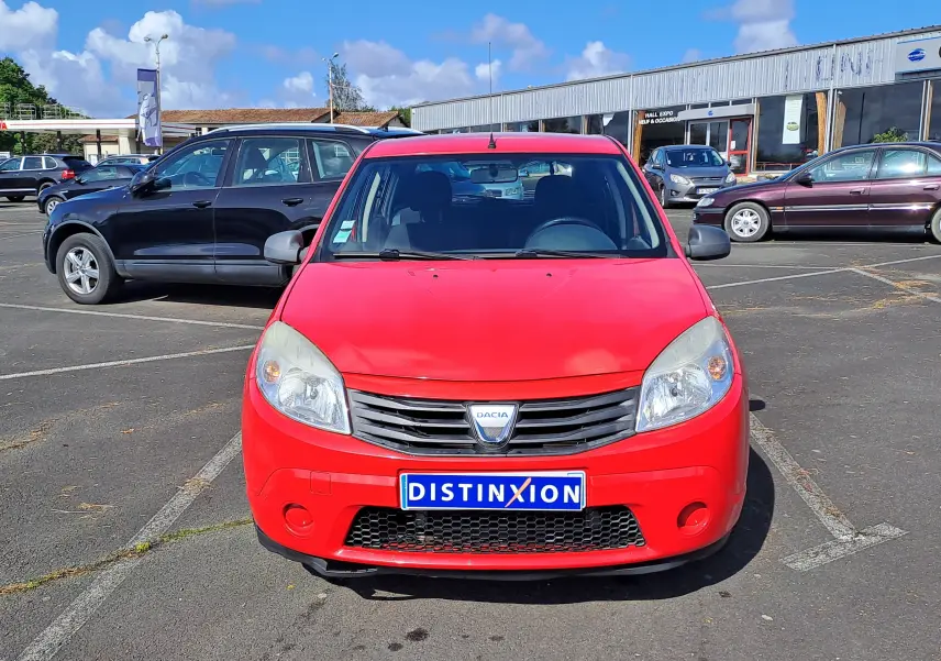 Vue de face d'une Dacia Sandero rouge de 2010 avec plaque Distinxion, stationnée sur un parking extérieur.