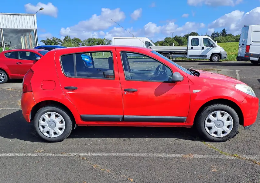 Profil côté gauche d'une Dacia Sandero rouge de 2010 avec jantes acier et poignées noires sur parking extérieur.