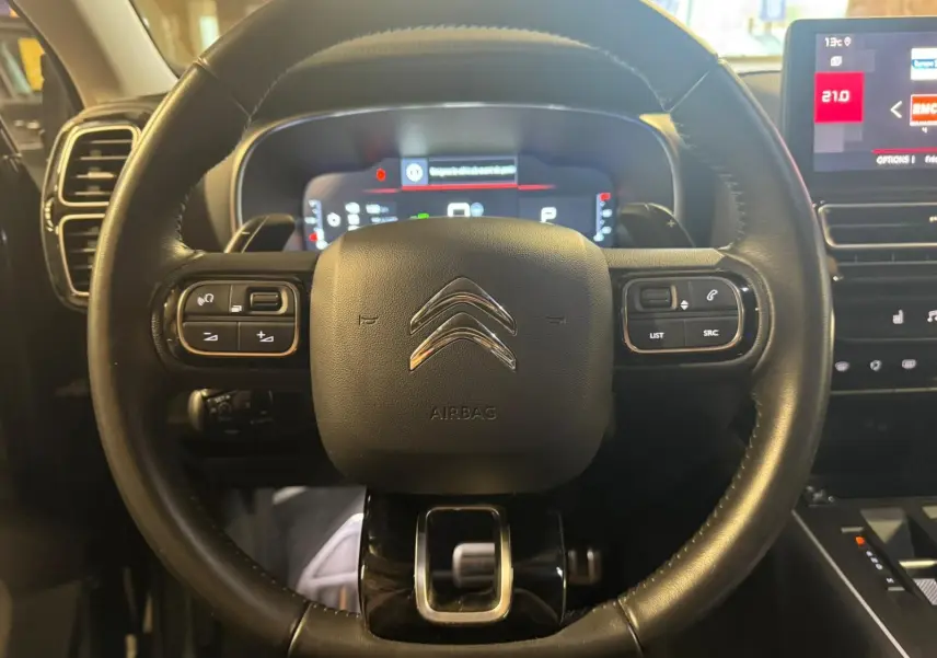 Gros plan sur les commandes noires du volant en cuir du Citroën C5 Aircross, avec écran numérique en arrière-plan.