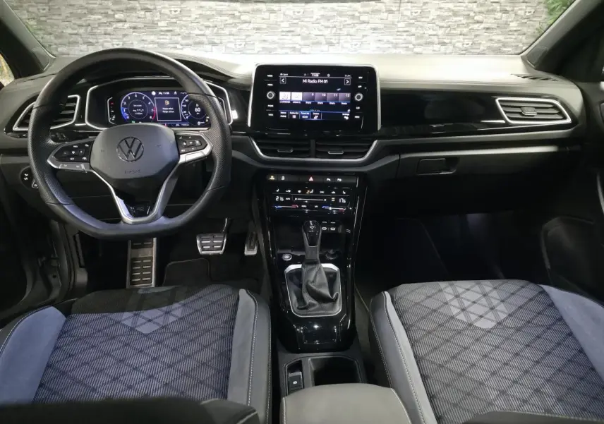 Vue intérieure avant du Volkswagen T-Roc 1.5 TSI R-Line 2025, avec tableau de bord digital et sièges tissu gris à motifs.