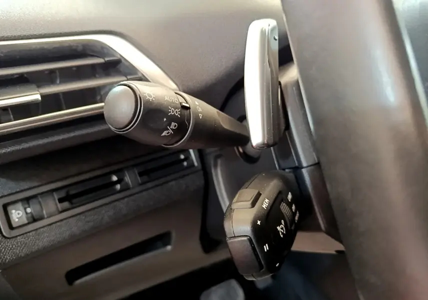 Gros plan sur la commande des phares et le volant cuir noir à gauche du tableau de bord du Peugeot 5008 gris clair.