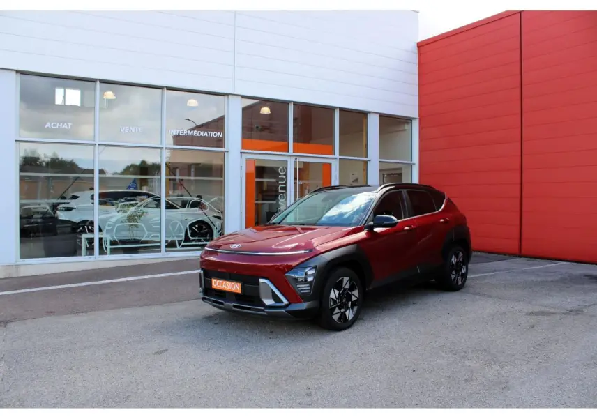 Hyundai Kona 2024 rouge Ultimate Metal avec toit noir, vu en 3/4 avant droit devant un bâtiment moderne.
