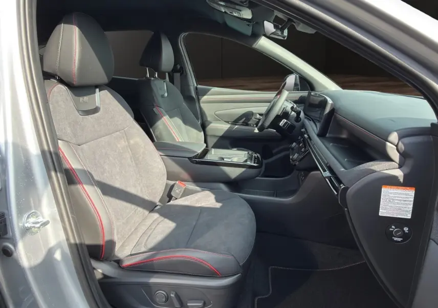 Vue intérieure côté conducteur du Hyundai Tucson 1.6 Hybrid N Line gris clair, sièges sport avec surpiqûres rouges et tableau de bord moderne.