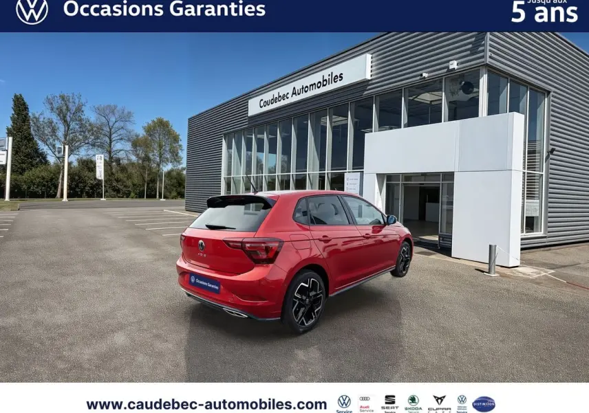 Vue 3/4 arrière droite d'une Volkswagen Polo rouge garée devant un bâtiment moderne de concession automobile.