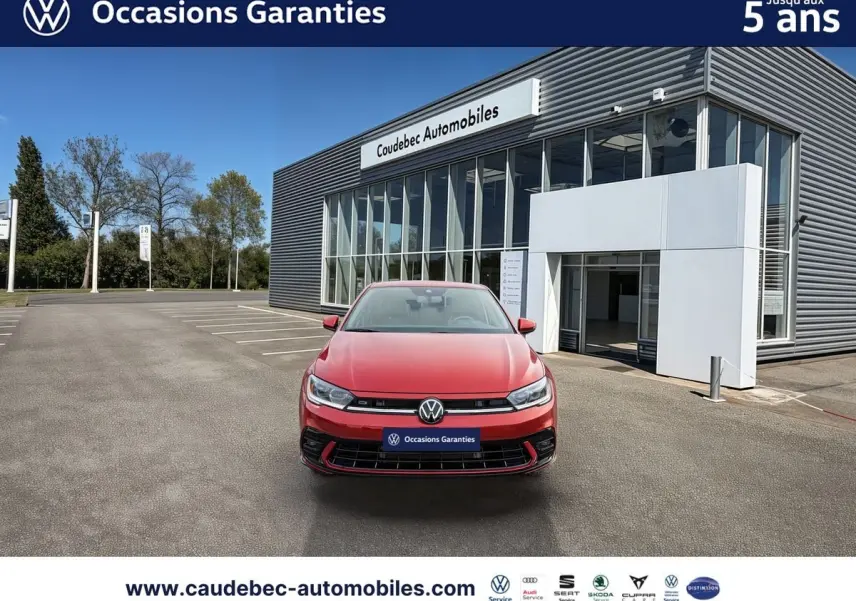 Vue avant d'une Volkswagen Polo rouge garée devant un bâtiment moderne de concession automobile Caudebec Automobiles.