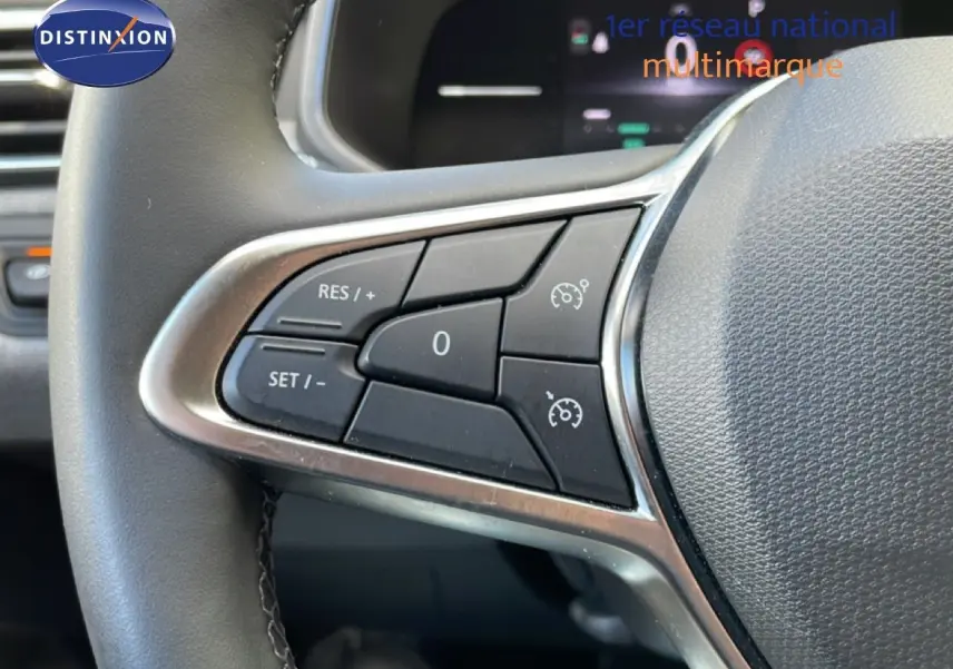 Gros plan sur les commandes au volant à gauche d’une Renault Clio gris schiste métal, avec affichage numérique en arrière-plan.