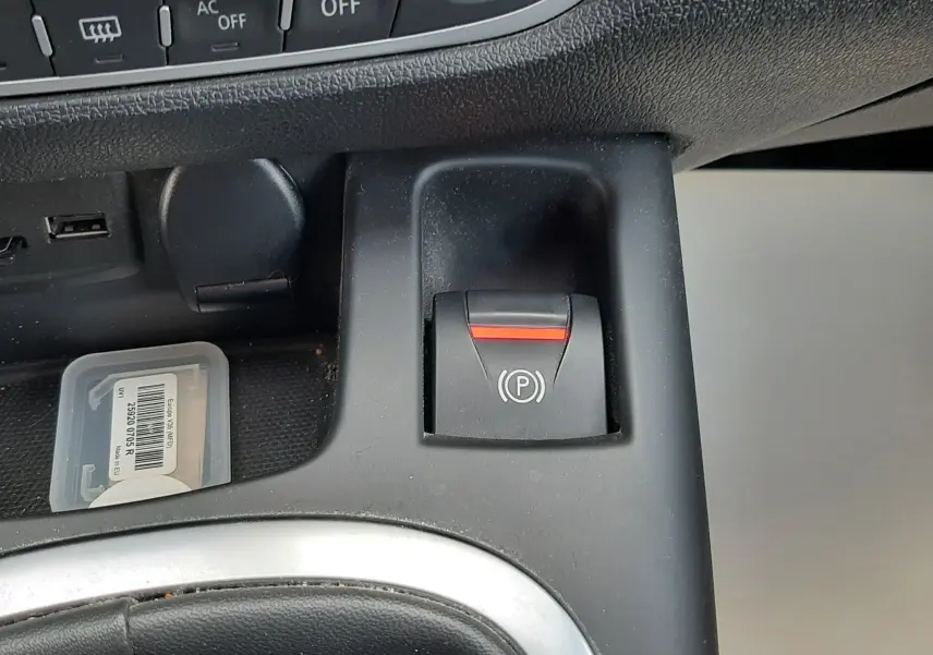 Gros plan sur le bouton de frein à main électrique noir avec témoin rouge, tableau de bord et console centrale du Renault Grand Scenic noir.