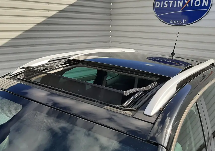 Toit ouvrant ouvert vu de dessus sur un Renault Grand Scenic noir avec barres de toit blanches, stationné près d'un mur.