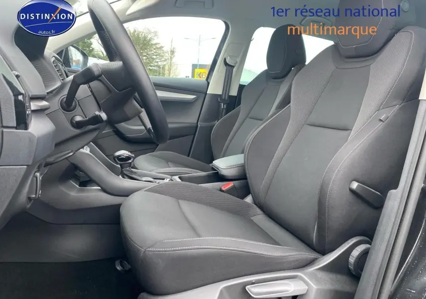 Vue intérieure côté conducteur du Skoda Karoq noir 2025, sièges tissu gris avec accoudoir central et volant cuir.