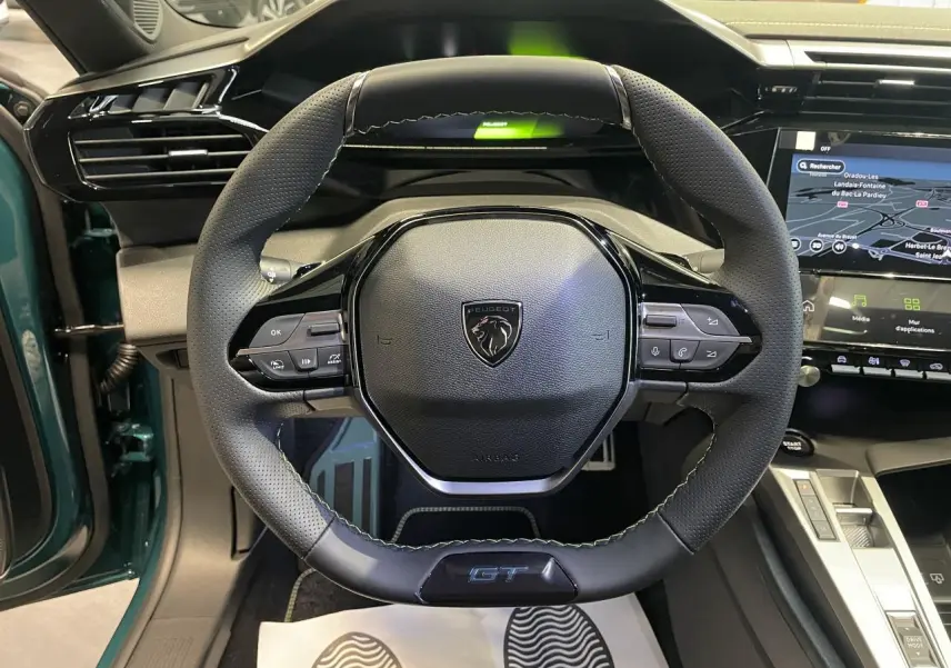 Vue rapprochée du volant GT en cuir noir avec surpiqûres vert Adamite dans l’habitacle bleu métallisé du Peugeot 308 2026.
