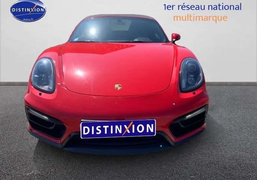 Vue frontale d'une Porsche Boxster rouge avec phares noirs et becquet arrière visible sur fond neutre.