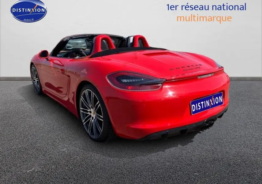 Vue 3/4 arrière droite d'une Porsche Boxster 981 rouge avec capote ouverte et jantes alu sport.