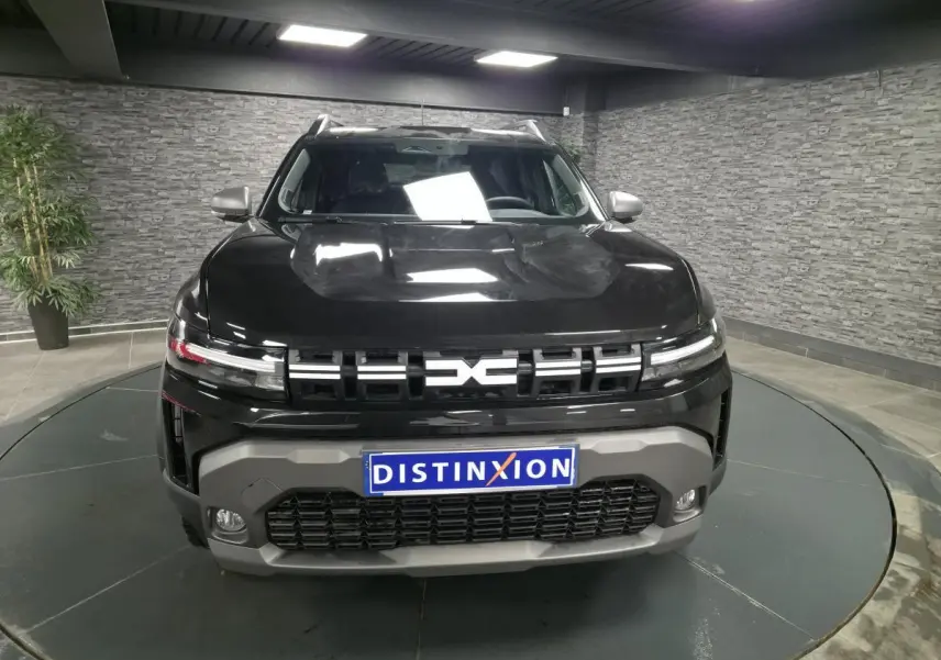 Vue avant d'un Dacia Duster 1.6 Hybrid noir nacré avec calandre moderne et plaque Distinxion en intérieur showroom.