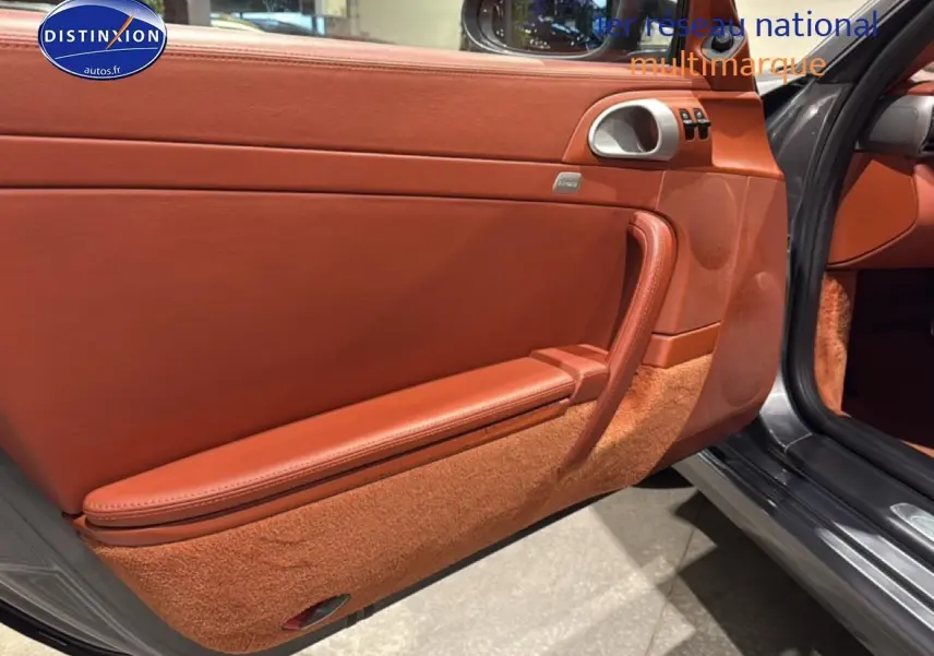 Vue intérieure côté gauche de la porte ouverte d’une Porsche 911 (997) Carrera PDK gris, avec garniture cuir marron et moquette assortie.