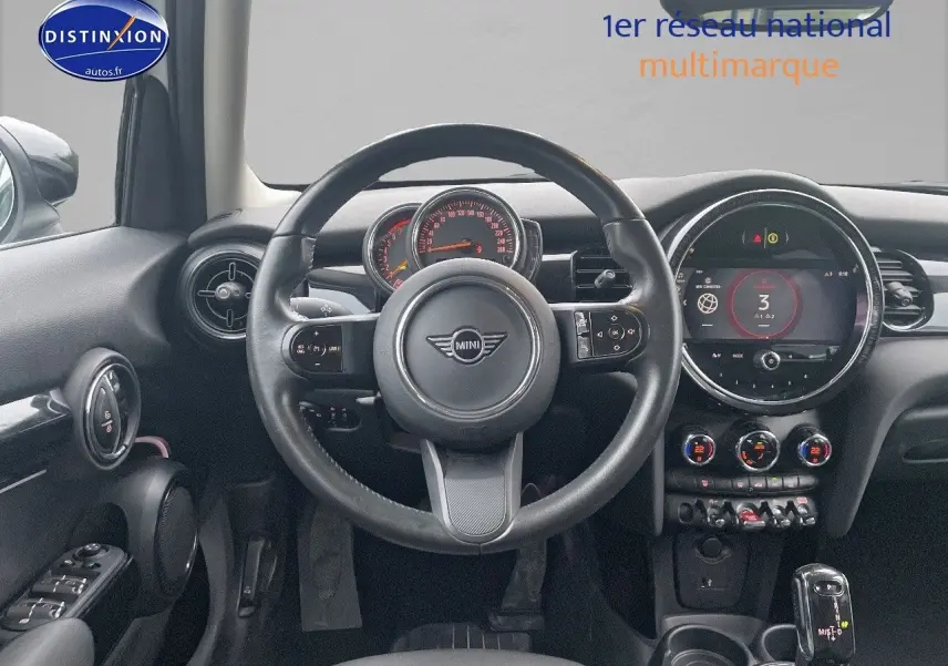 Vue intérieure centrée sur le volant et le tableau de bord d'une MINI blanche nacrée 2023 avec écran numérique et commandes modernes.