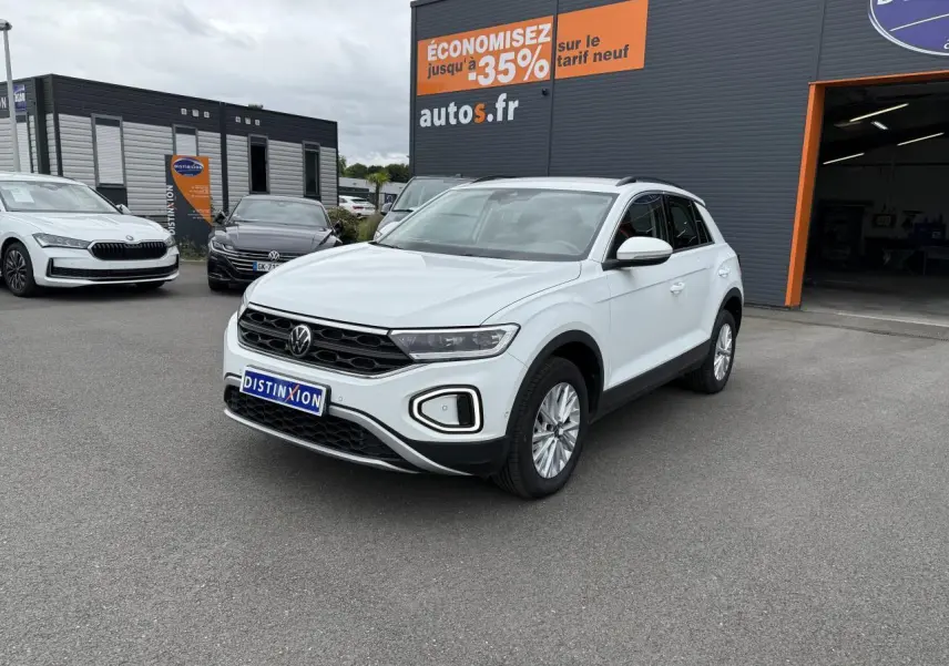 Volkswagen T-Roc blanc pur vu en 3/4 avant droit sur parking devant un garage avec panneau promotionnel orange.
