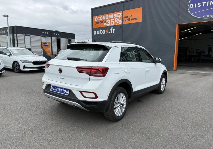 Vue 3/4 arrière droite d'un Volkswagen T-Roc blanc pur garé devant un concessionnaire avec panneaux publicitaires visibles.