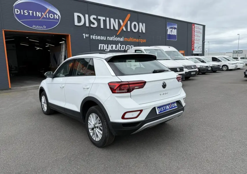 Volkswagen T-Roc blanc pur vu en 3/4 arrière droit, garé devant un bâtiment avec plusieurs véhicules blancs alignés.