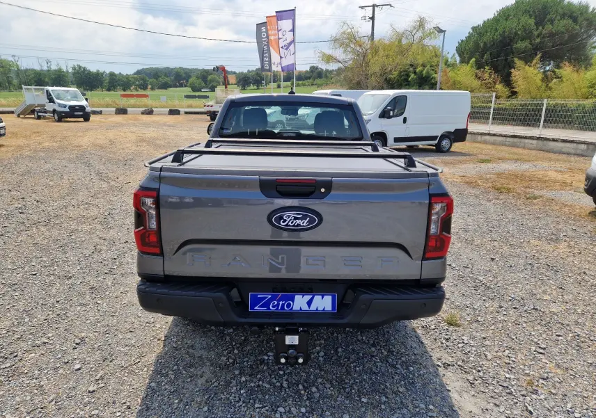 Vue arrière d'un Ford Ranger Super Cabine gris carbone avec rideau de plateau rigide et attelage visible sur un terrain gravillonné.