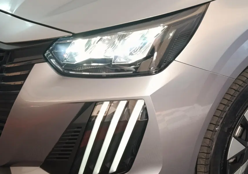 Gros plan sur l'avant droit gris clair de la Peugeot 208 électrique 2024, mettant en valeur les phares LED et les feux diurnes verticaux.