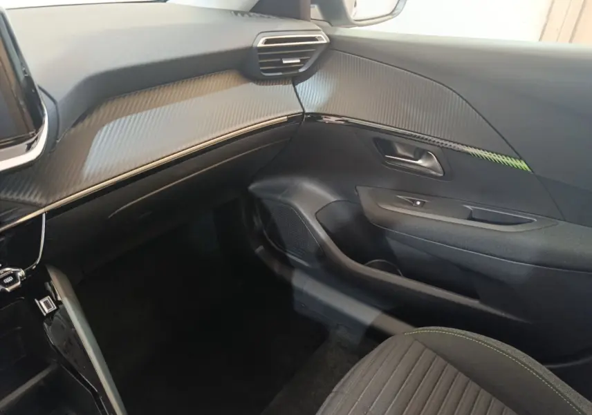 Intérieur côté passager du Peugeot 208 électrique 2024, tableau de bord noir avec insert effet carbone et surpiqûres vertes sur siège.