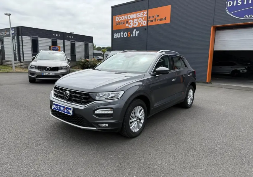 Volkswagen T-Roc gris Indium Metal en 3/4 avant droit, stationné devant un garage avec panneaux publicitaires.